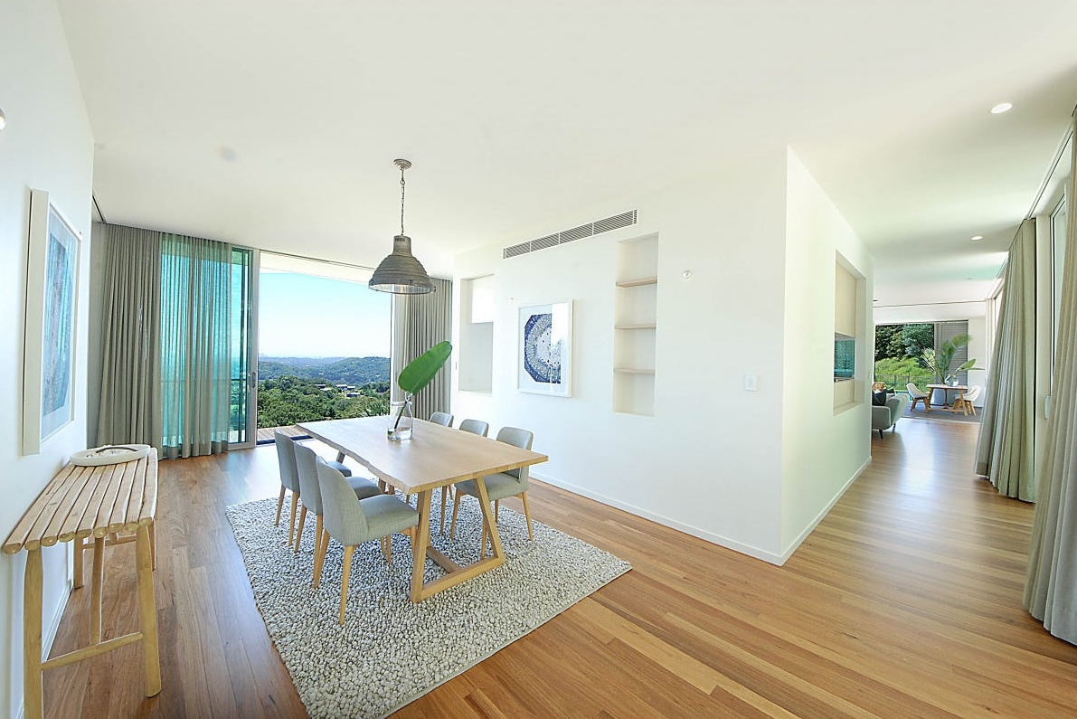 Dining room at Brocks Road with a timber table, clean white walls and a view through glass doors to the hinterland, with no visible technology.