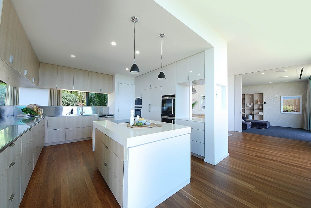 Kitchen at Brocks Road with a white island, pale timber cabinetry, black pendant lights and clean ceilings with no visible speakers.