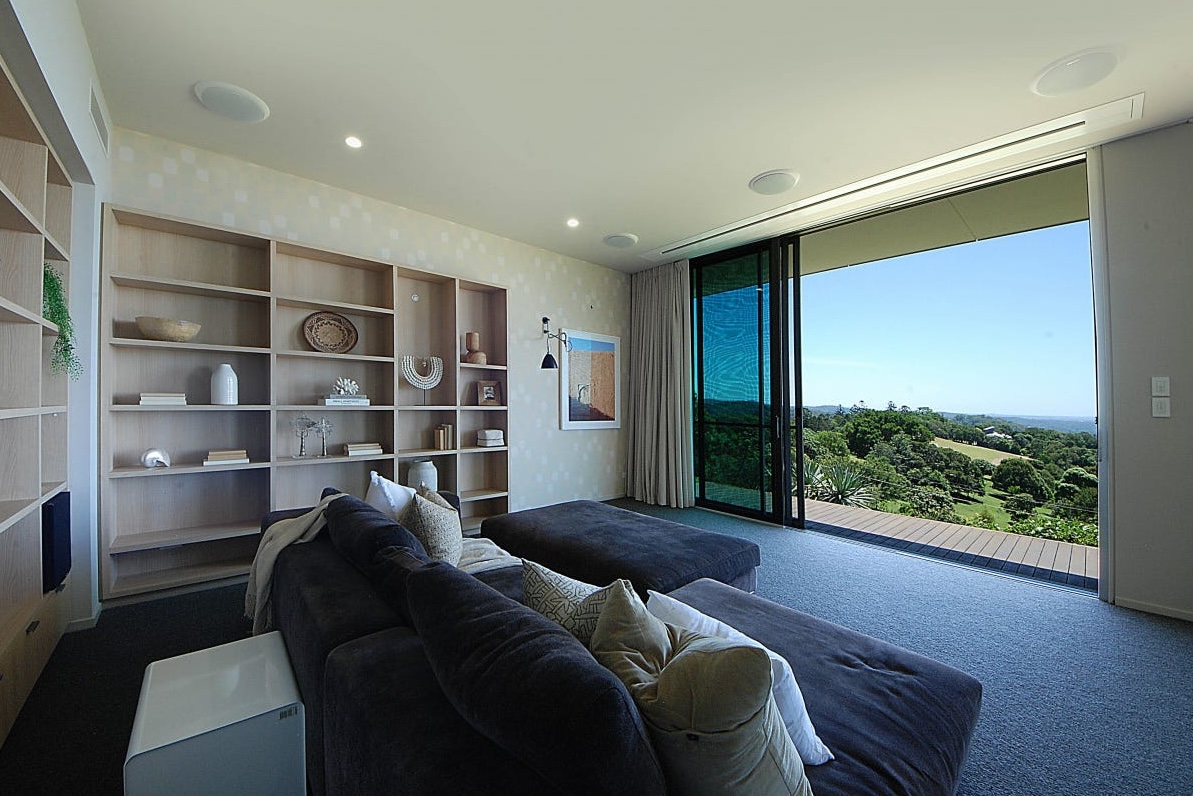 Media room at Brocks Road with a deep sofa, built-in timber shelving, flush in-ceiling speakers and glass doors open to the hinterland, with the screen concealed in the ceiling.