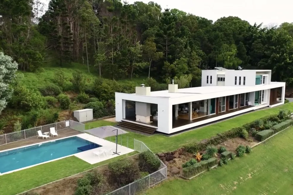 Aerial view of the Brocks Road home, a long white residence with a pool, set against a forested hillside.