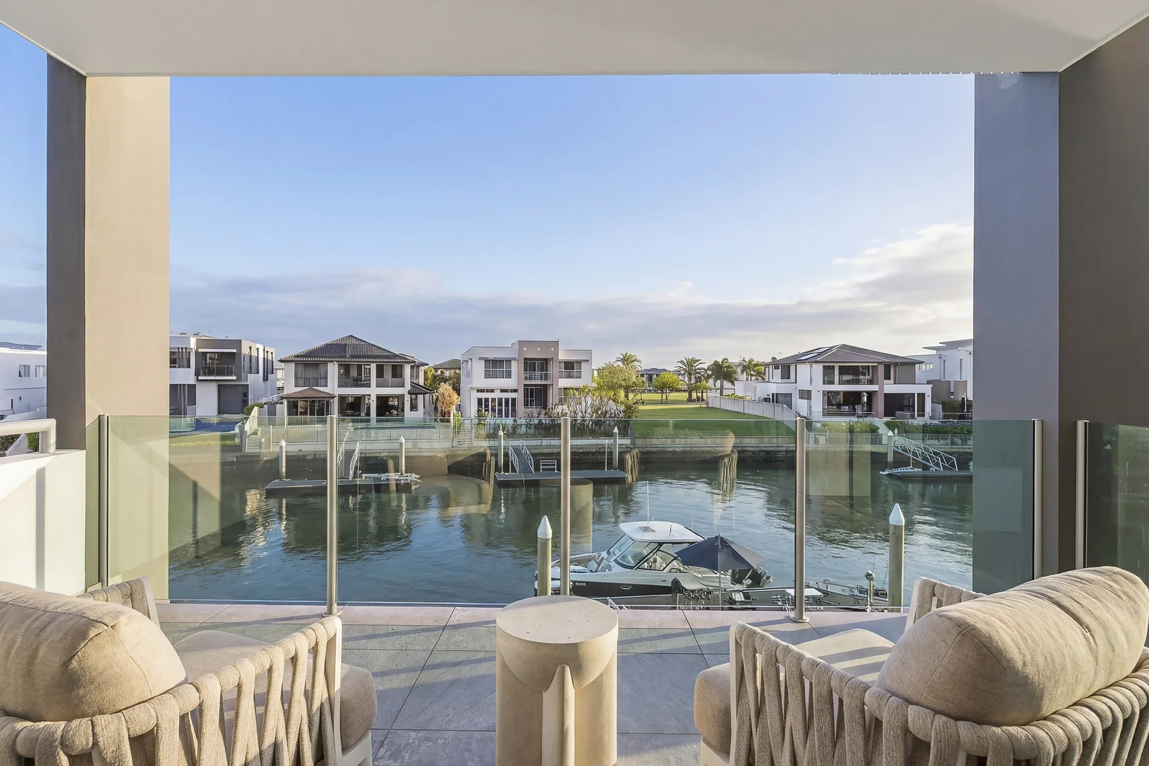 Covered balcony at Knightsbridge with two woven armchairs and a glass balustrade, looking over the canal to a moored boat and homes across the water.