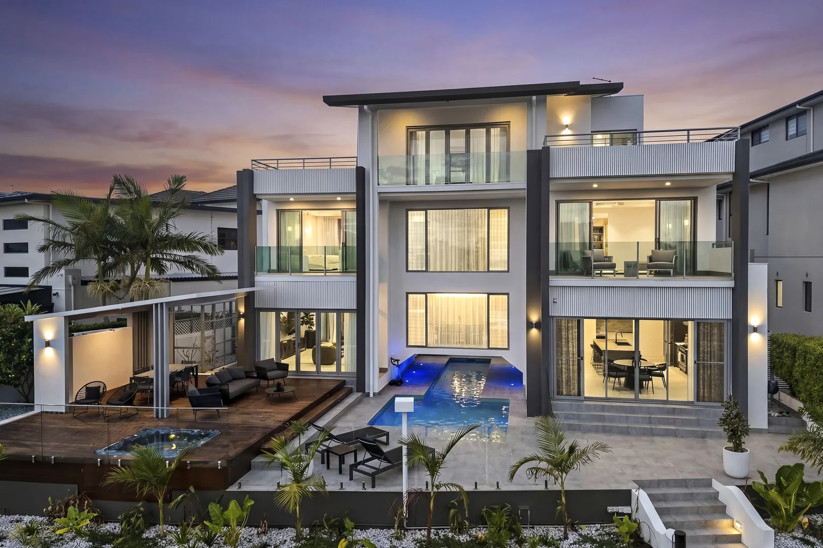 The waterfront side of the Knightsbridge home at dusk, three storeys lit from within, with a pool and timber deck.