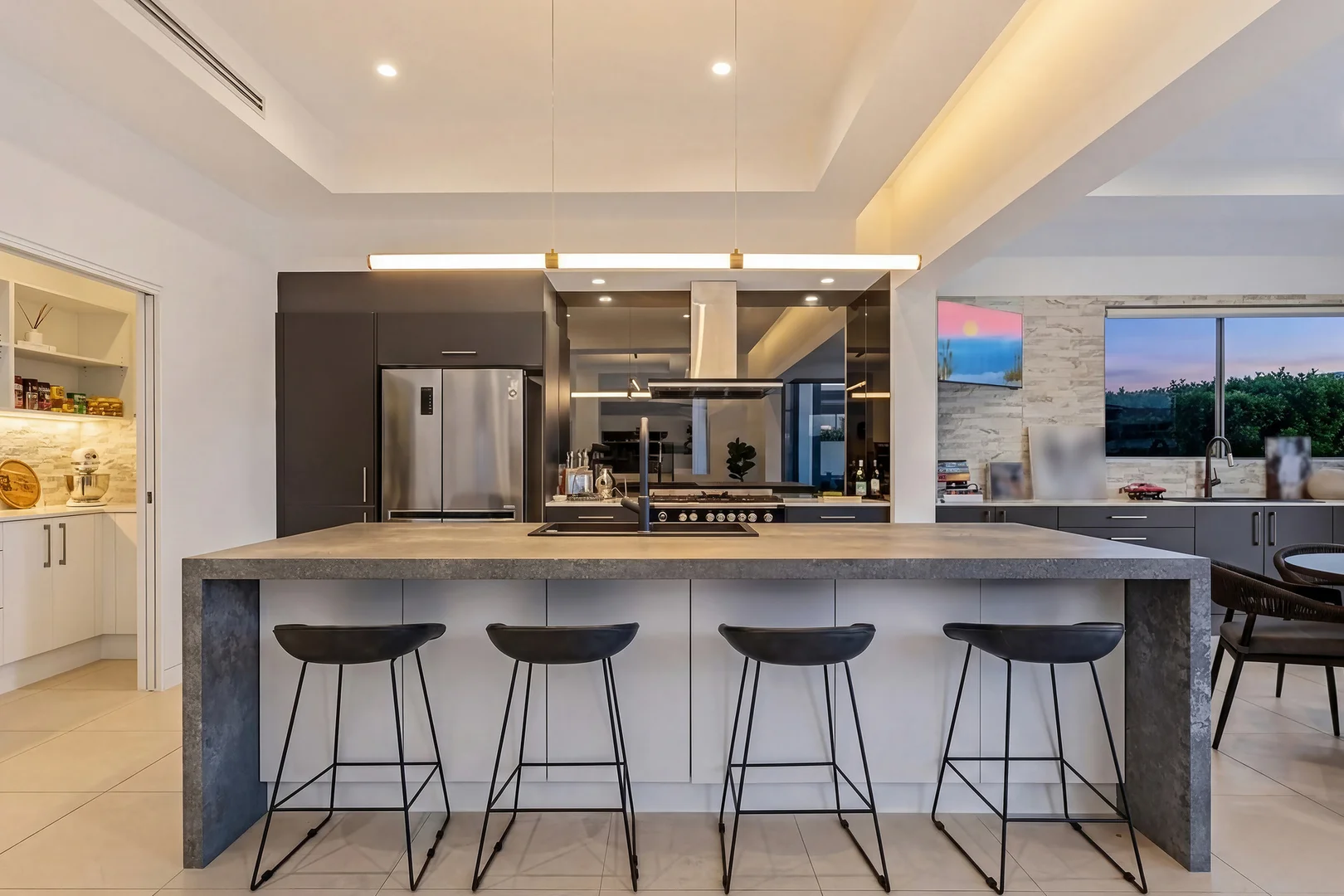 Kitchen with a concrete-look island and four black stools, with a television on the stone wall at the far right.
