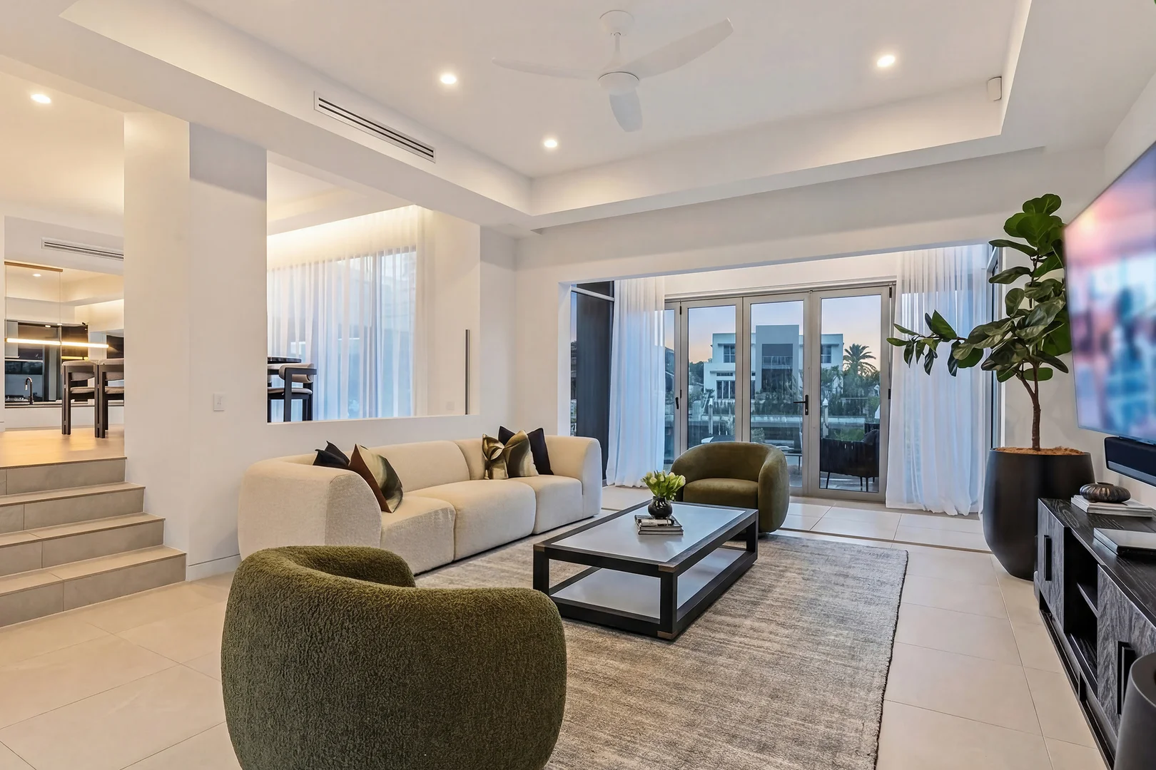 Living room with a cream sofa, two green armchairs and a wall-mounted television with soundbar, opening to the waterfront.