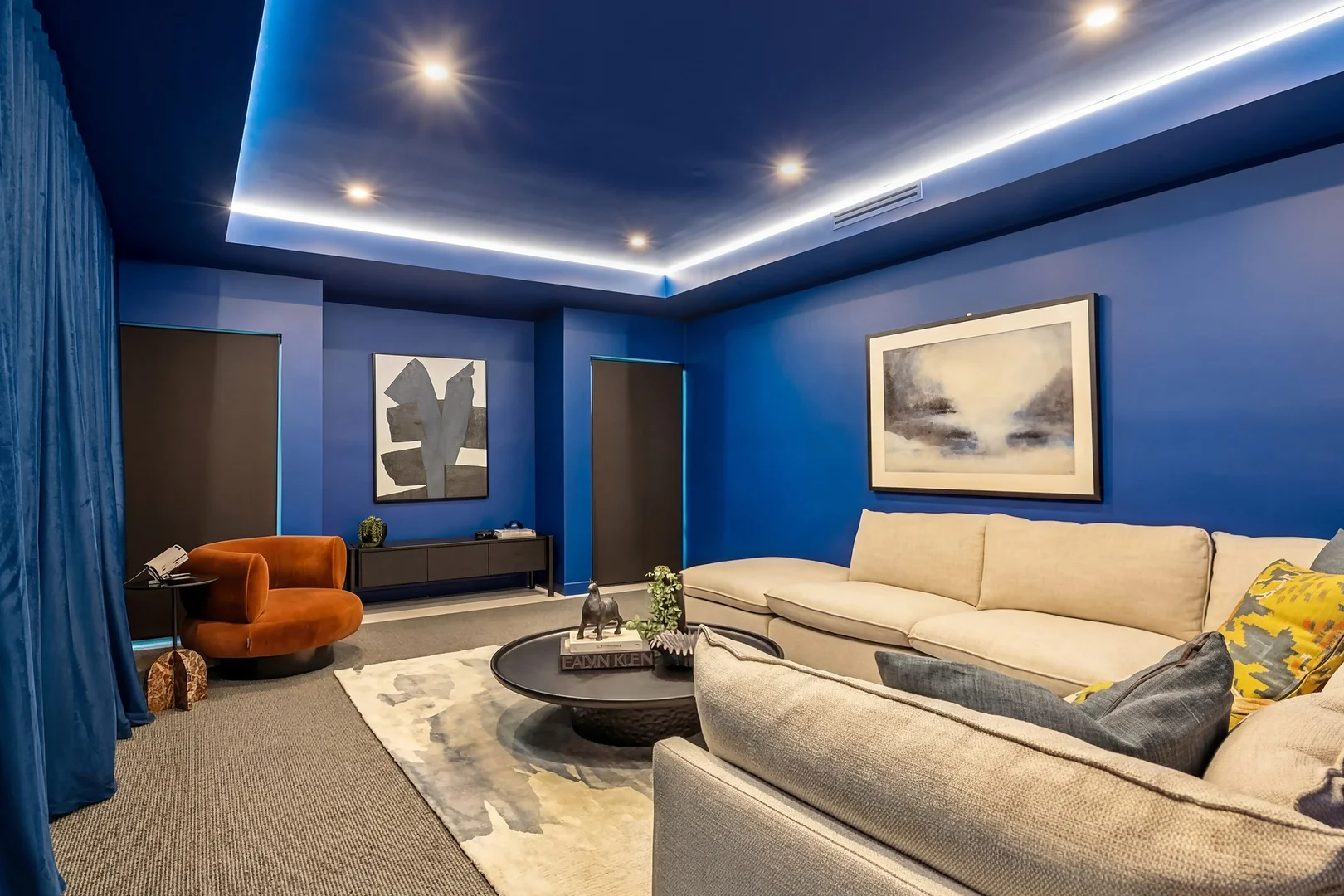 Media room with deep blue walls and curtains, a cream sectional sofa, an orange armchair and a backlit ceiling tray.