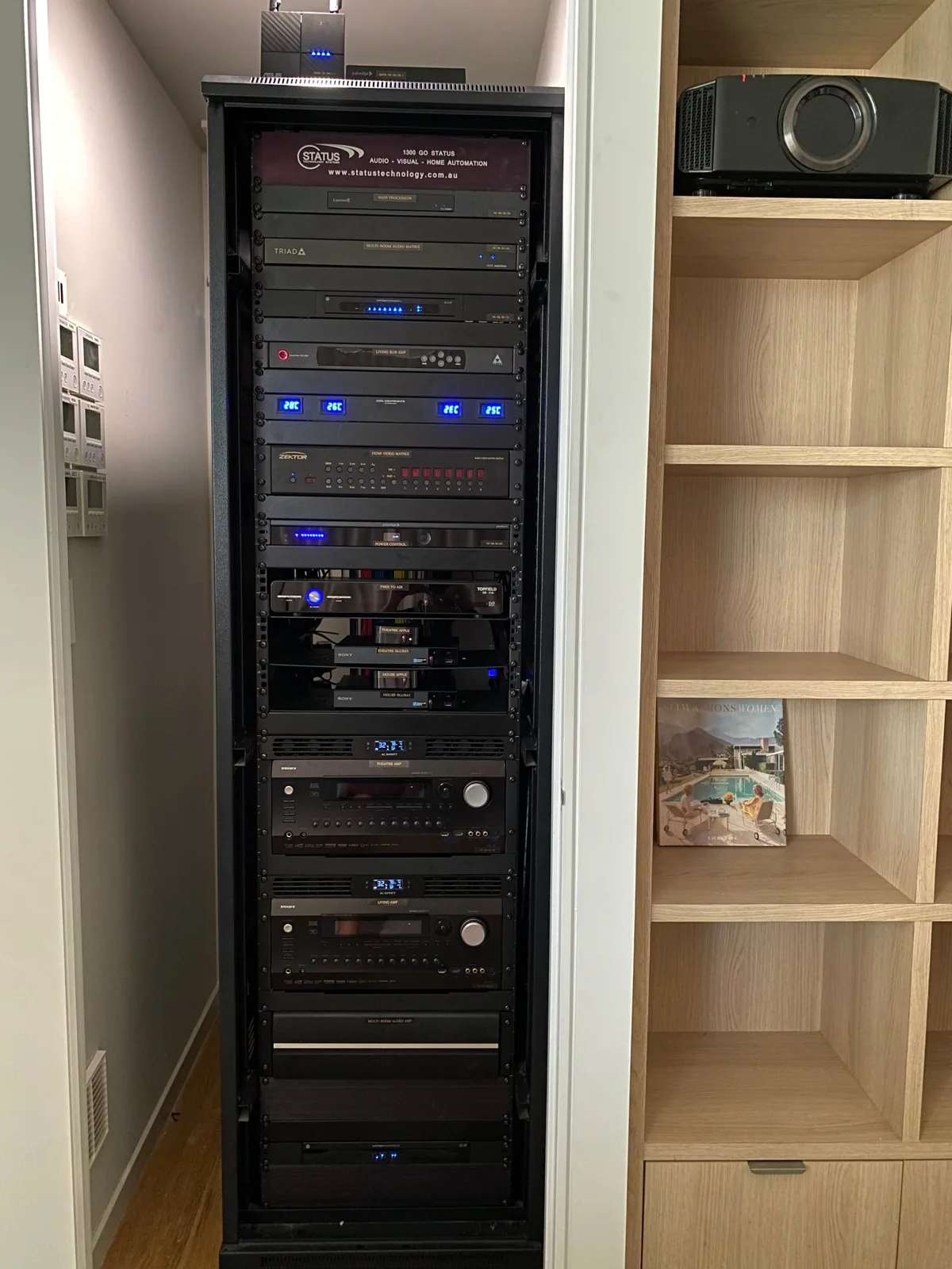 Black AV equipment rack at Brocks Road with labelled components, blue temperature displays and cooling fans, beside timber shelving with a JVC projector.