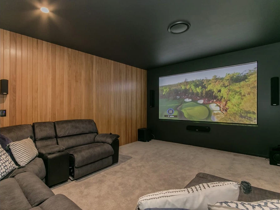 Dark home theatre with a large projection screen, wall-mounted speakers, timber wall lining and reclining sofas.