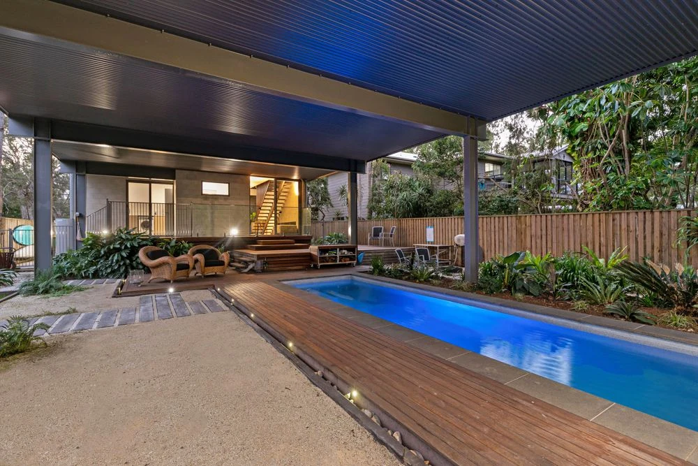 Illuminated sheltered pool with timber decking, planting and outdoor seating at dusk.